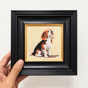 May include: A small square painting of a beagle dog with brown, black, and white fur, sitting and looking upward. The artwork is encased in a thick, matte black frame with a thin gold inner border, presented against a plain white background.