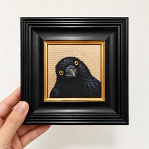 Pode incluir: Uma pequena pintura quadrada de um corvo preto com olhos amarelos brilhantes, sobre um fundo bege texturizado. A obra está emoldurada numa moldura preta espessa e brilhante com uma fina borda interior dourada, criando um contraste nítido.