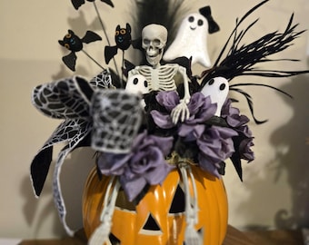 Puede incluir: Un centro de mesa decorativo de calabaza naranja con una figura de esqueleto central, adornos de fantasmas blancos, rosas de tela moradas, detalles de plumas negras y cinta con patrón de telaraña en blanco y negro.