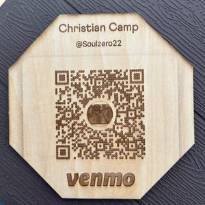 May include: An octagonal light-coloured wooden sign featuring a laser-engraved QR code in the centre. The text "Christian Camp", "@Soulzero22", and "venmo" are engraved in dark brown lettering above and below the central QR code graphic.