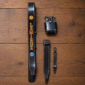 May include: A black leather radio strap set featuring a long strap with the text LEMIEUX in yellow and red, a red and yellow FD emblem, and a blue star of life symbol. The set includes a black leather radio holder, a short strap, and a connector.