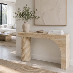 May include: A beige travertine console table featuring a single spiraled cylindrical leg on the left and a smooth arched support on the right. The table surface holds a textured ceramic vase with green foliage, a small bowl, and two stacked books.