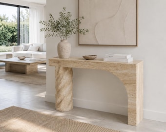 Può includere: Un tavolo console in travertino beige caratterizzato da una singola gamba cilindrica a spirale sulla sinistra e un supporto ad arco liscio sulla destra. Il piano del tavolo ospita un vaso in ceramica testurizzata con fogliame verde, una piccola ciotola e due libri impilati.