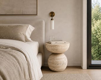 Può includere: Un tavolino in travertino beige con base sferica e piano circolare, posizionato accanto a un letto. Il tavolo sostiene un bicchiere di vetro trasparente e un libro con la scritta "at 100" sulla copertina, sotto una lampada da parete in ottone e bianco.