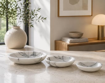 Può includere: Un set di tre ciotole circolari in marmo bianco con venature grigie, disposte in ordine decrescente di dimensioni su una superficie in pietra chiara. Ogni ciotola presenta una finitura liscia e lucida e un design a base arrotondata.