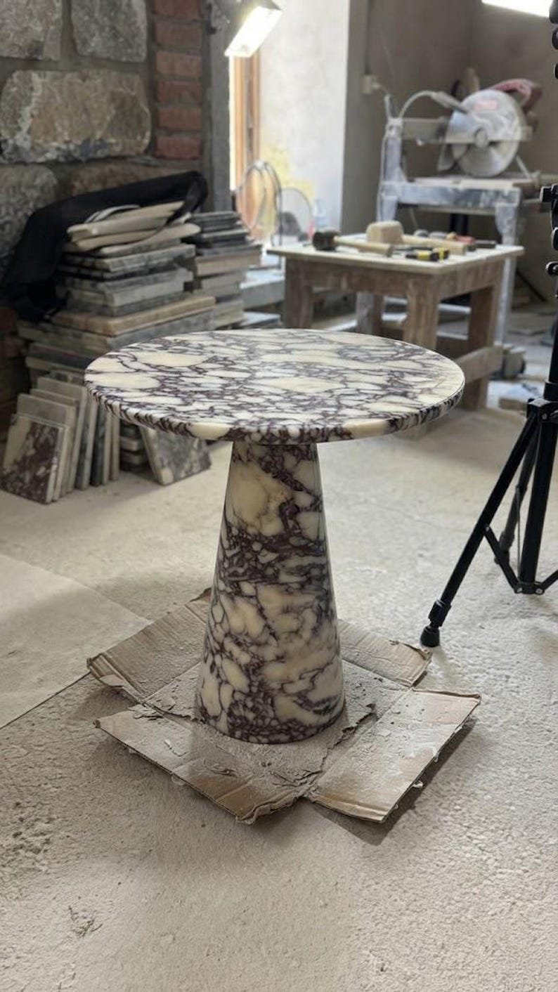 May include: A round side table crafted from white and dark purple veined marble. The table features a circular top supported by a single, wide, conical pedestal base that tapers toward the top, showcasing a consistent natural stone pattern throughout.