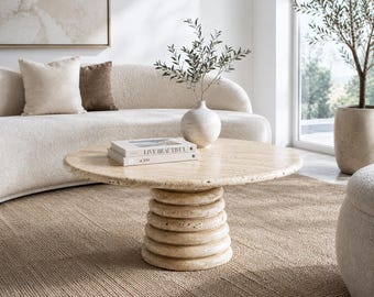 May include: A round beige travertine coffee table featuring a thick, textured stone top and a tiered, circular pedestal base. Two stacked books are placed on the surface, with the top book clearly displaying the text LIVE BEAUTIFUL.