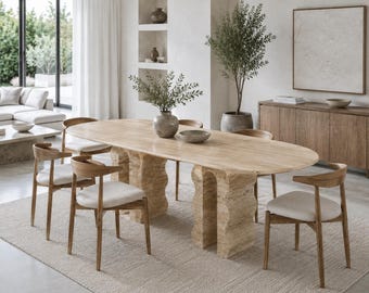 Può includere: Un tavolo da pranzo ovale con un piano in pietra beige liscia e due basi a piedistallo in pietra spesse, strutturate e dai bordi ondulati. Il tavolo è circondato da quattro sedie in legno con schienali curvi e cuscini color bianco sporco.