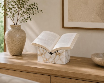Può includere: Un leggio in marmo bianco con venature dorate, caratterizzato da un design a forma di V che sostiene un libro aperto con pagine di testo visibili. Il supporto è rettangolare con una finitura liscia e lucida, offrendo una base stabile.