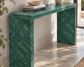 Può includere: Un tavolo console a cascata realizzato in marmo verde scuro con venature bianche prominenti. Il tavolo presenta una superficie liscia e lucida e un design continuo a forma di U. Due libri intitolati The Kinfolk Home e At Home sono appoggiati sopra.