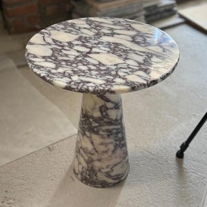 May include: A round side table crafted from white and dark purple veined marble. The table features a circular top surface supported by a single, solid, tapered conical base that matches the marble pattern of the tabletop throughout the piece.