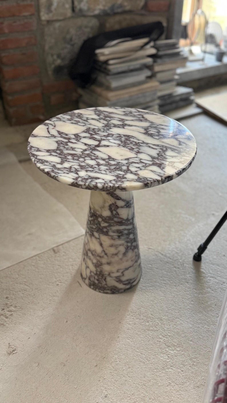 May include: A round side table crafted from white and dark purple veined marble. The table features a circular top surface supported by a single, solid, tapered conical base that matches the marble pattern of the tabletop throughout the piece.