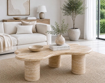 May include: A beige, organic-shaped coffee table made of travertine stone with three cylindrical legs. The table surface features a natural porous texture and holds a small bowl, two stacked books with the text LIVE BEAUTIFUL, and a vase.