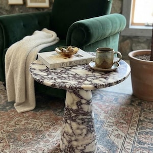May include: A round side table made of white and purple veined marble with a pedestal base. On the table sits a book titled Live Beautiful, a small amber glass bowl containing dried botanicals, and a speckled ceramic mug on a matching saucer.