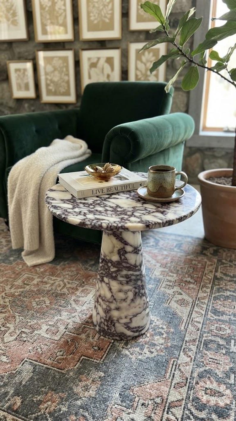 May include: A round side table made of white and purple veined marble with a pedestal base. On the table sits a book titled Live Beautiful, a small amber glass bowl containing dried botanicals, and a speckled ceramic mug on a matching saucer.