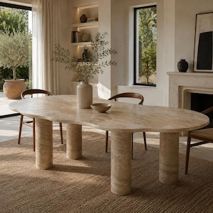 Puede incluir: Una mesa de comedor de forma orgánica hecha de piedra travertino beige, con una superficie de tablero lisa e irregular sostenida por cuatro patas cilíndricas gruesas. La mesa se acompaña de tres sillas de madera con asientos trenzados.
