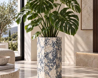 Può includere: Una grande pianta di Monstera deliciosa con ampie foglie verdi divise, inserita in un alto vaso esagonale caratterizzato da un motivo in pietra maculata bianca e blu scuro, con finitura liscia e bordi geometrici netti.