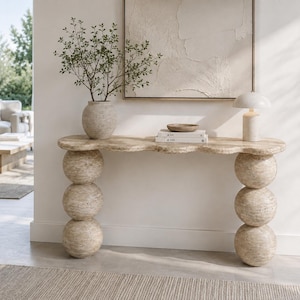 Op de afbeelding: Een beige travertijn sidetable met een lang blad met geschulpte randen, ondersteund door twee verticale kolommen die elk bestaan uit drie gestapelde bollen. Het natuurstenen oppervlak heeft een poreuze, getextureerde afwerking in aardetinten.