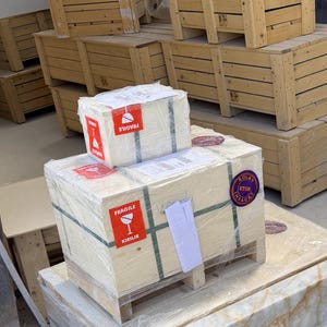 May include: Two rectangular wooden crates stacked on a pallet, wrapped in clear plastic film. The crates feature red and white fragile labels with the text FRAGILE and KIRILIR, along with a purple circular sticker labeled KOLAY ETGB IHRACAT.