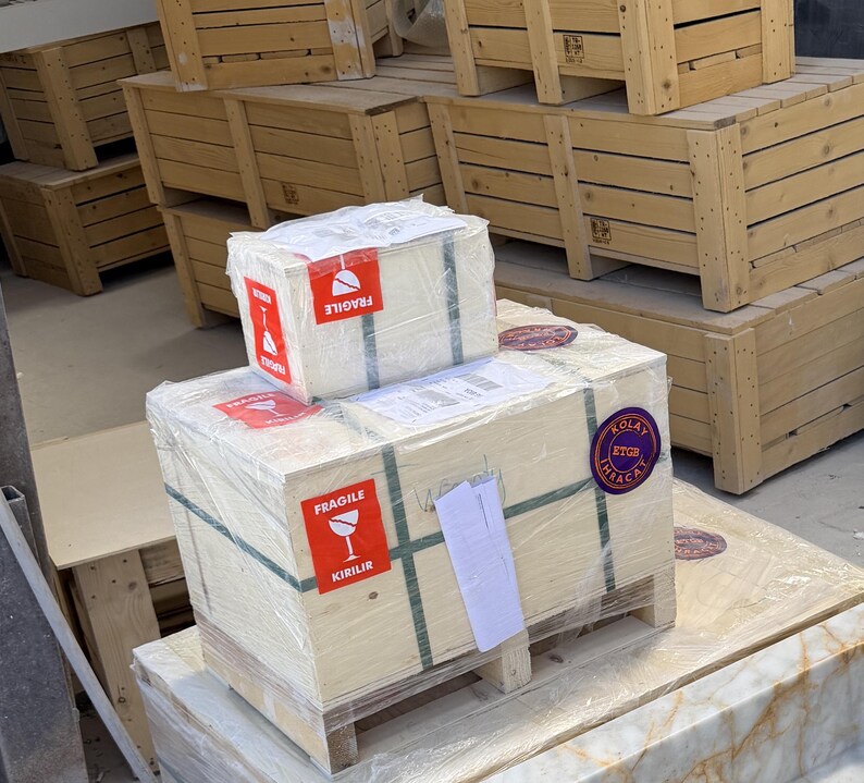 May include: Two rectangular wooden crates stacked on a pallet, wrapped in clear plastic film. The crates feature red and white fragile labels with the text FRAGILE and KIRILIR, along with a purple circular sticker labeled KOLAY ETGB IHRACAT.