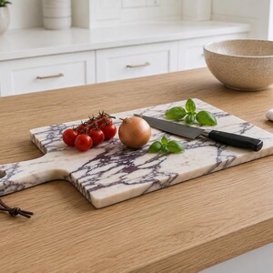 May include: A rectangular white marble cutting board with dark purple veining, featuring a handle with a brown leather cord. On the board are a cluster of red cherry tomatoes, a single yellow onion, a few basil leaves, and a metal kitchen knife.