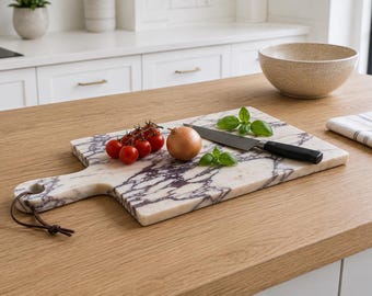 Può includere: Un tagliere rettangolare in marmo bianco con venature viola scuro, dotato di un manico con un cordino in pelle marrone. Sul tagliere si trovano un grappolo di pomodorini rossi, una cipolla gialla, foglie di basilico e un coltello da cucina.