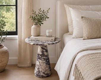 Può includere: Un tavolino rotondo con base conica, realizzato in marmo bianco con venature viola scuro. Il piano del tavolo ospita un vaso in ceramica beige con fogliame verde, un libro bianco intitolato AD at 100 e una piccola candela in vetro.