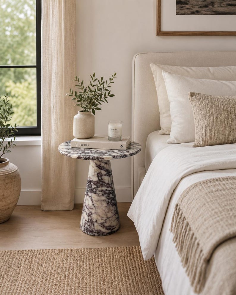 May include: A round side table with a conical base, crafted from white marble featuring prominent dark purple veining. The table surface holds a beige ceramic vase with green foliage, a white book titled AD at 100, and a small glass candle.