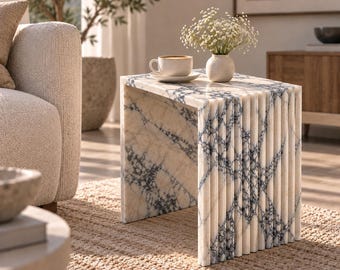 Può includere: Un tavolino rettangolare in marmo bianco con venature blu scuro prominenti. Il tavolo presenta un design a cascata con dettagli scanalati verticali lungo il pannello laterale. Sopra sono posizionati una tazza in ceramica bianca e un piccolo vaso.