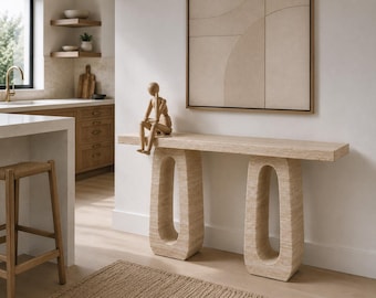 Può includere: Un tavolo consolle in travertino beige con piano rettangolare e due basi cave di forma ovale. Un piccolo manichino articolato in legno è seduto sul bordo sinistro della superficie del tavolo. La pietra presenta una texture naturale e porosa.