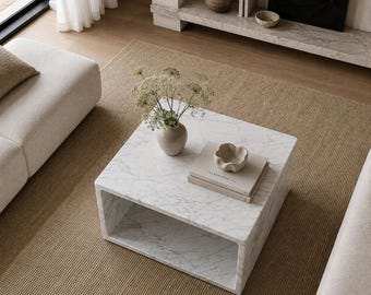 Può includere: Un tavolino quadrato in marmo bianco con un ripiano centrale cavo. Sul piano del tavolo si trovano un vaso in ceramica beige con fiori secchi, una piccola ciotola in ceramica beige smerlata e due libri impilati con copertine chiare.