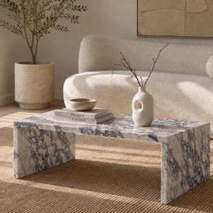 Puede incluir: Una mesa de centro rectangular hecha de mármol blanco con vetas azul oscuro prominentes. La mesa presenta un diseño de borde en cascada con dos paneles laterales sólidos. Sobre la superficie hay dos libros apilados, un cuenco y un jarrón de cerámica.