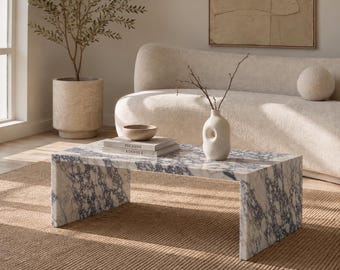 May include: A rectangular coffee table made of white marble with prominent dark blue veining. The table features a waterfall edge design with two solid side panels. On the surface sit two stacked books, a small bowl, and a white ceramic vase.
