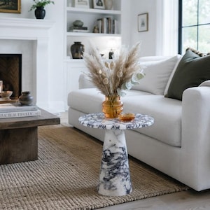 May include: A round side table featuring a white and black marbled stone finish with a tapered pedestal base. An amber-colored glass vase filled with dried pampas grass and seed pods sits on the tabletop, accompanied by a small amber glass dish.