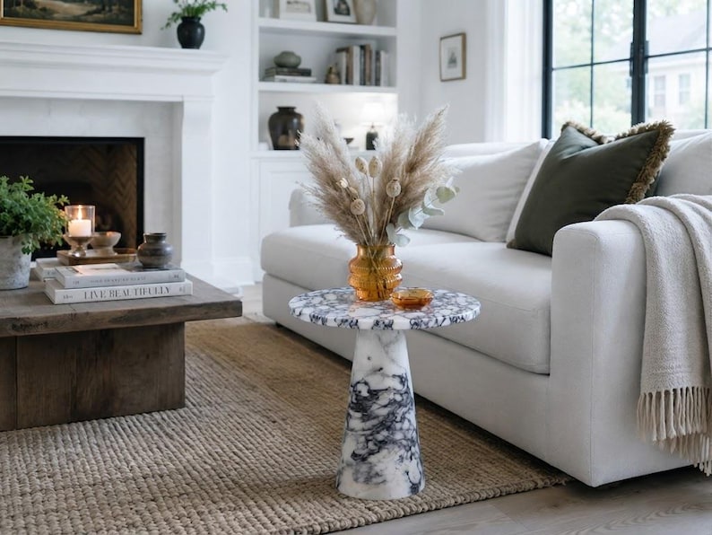 May include: A round side table featuring a white and black marbled stone finish with a tapered pedestal base. An amber-colored glass vase filled with dried pampas grass and seed pods sits on the tabletop, accompanied by a small amber glass dish.