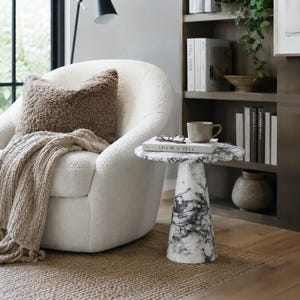 May include: A white marble side table with a conical base and a circular top, featuring black veining throughout. The table is positioned next to a white textured armchair draped with a beige knit throw and a brown fuzzy pillow, with a book on top.