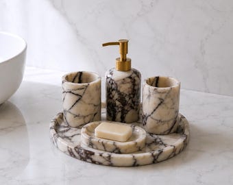 Può includere: Un set di accessori da bagno realizzato in marmo color crema con venature marrone scuro. Il set comprende un dispenser di sapone cilindrico con erogatore dorato, due bicchieri cilindrici abbinati, un portasapone ovale e un vassoio circolare.