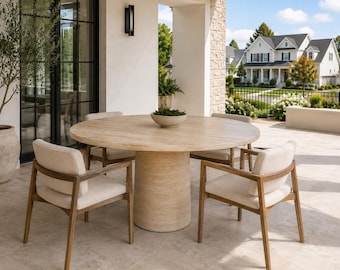 Può includere: Un tavolo da pranzo rotondo in pietra beige con base a piedistallo centrale e quattro poltrone in legno con sedute e schienali imbottiti color crema chiaro. Una piccola ciotola poco profonda è posizionata al centro su un patio in pietra.