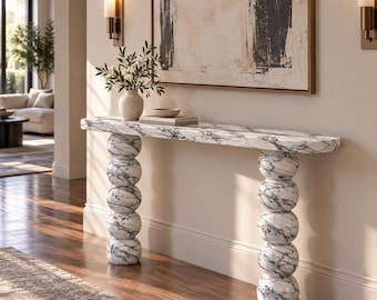 Può includere: Un tavolo console in marmo bianco con venature nere, caratterizzato da un lungo piano rettangolare liscio sostenuto da due colonne verticali composte da sfere di marmo impilate. Il tavolo è posizionato contro una parete sotto un grande dipinto.