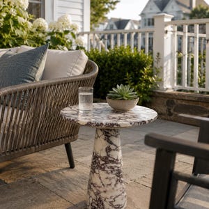 May include: A round side table featuring a white and dark purple marble pattern with a conical base. The table surface holds a clear glass of water and a small, light-colored bowl containing a green succulent plant, positioned outdoors.