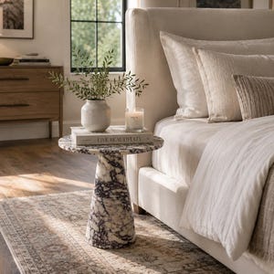 May include: A round side table featuring a white and dark purple marble pattern with a conical base. The table surface holds a white ceramic vase with green foliage, a glass candle, and a book with the text LIVE BEAUTIFULLY on the cover.