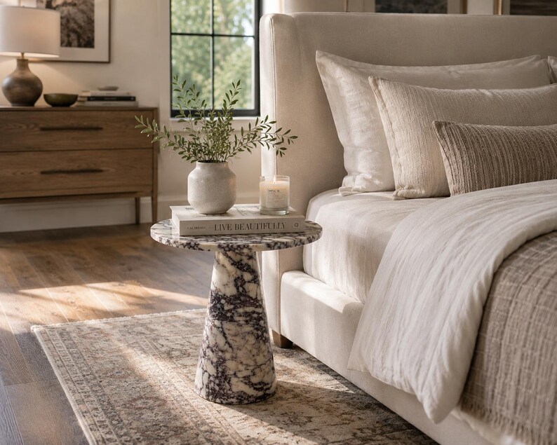 May include: A round side table featuring a white and dark purple marble pattern with a conical base. The table surface holds a white ceramic vase with green foliage, a glass candle, and a book with the text LIVE BEAUTIFULLY on the cover.