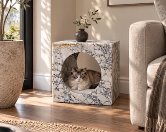 Può includere: Un tavolino quadrato con motivo in marmo bianco e grigio, caratterizzato da un'ampia apertura circolare centrale. Un gatto a pelo lungo dal manto marrone e bianco riposa all'interno dell'apertura circolare su un morbido cuscino chiaro.