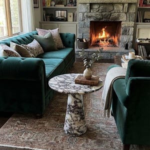 May include: A living room featuring a dark green tufted velvet sofa with patterned throw pillows, a round white and gray marble pedestal coffee table, a matching green velvet armchair with a light gray throw blanket, and a stone fireplace.