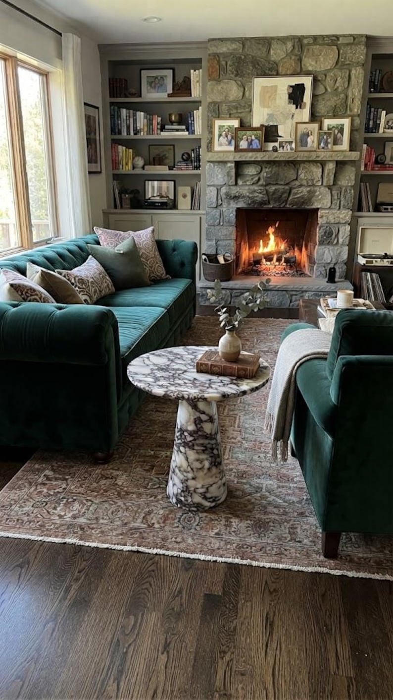 May include: A living room featuring a dark green tufted velvet sofa with patterned throw pillows, a round white and gray marble pedestal coffee table, a matching green velvet armchair with a light gray throw blanket, and a stone fireplace.