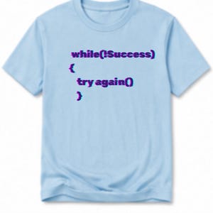 May include: A light blue short-sleeved t-shirt featuring the dark purple and blue glitch-effect text: while(!Success) { try again() }. The graphic is centered on the chest of the shirt, which has a crew neckline and a standard straight hem.