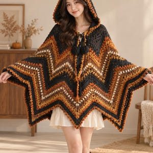 Può includere: Un poncho all'uncinetto con cappuccio caratterizzato da un motivo a chevron in nero, arancione bruciato, giallo senape e crema. Il capo include un cappuccio, una chiusura con coulisse con due pon-pon neri sul collo e un orlo a stella dentellato.