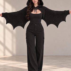 May include: A black crochet shrug featuring a hood with cat-like ears and bat-wing shaped sleeves. The garment is worn over a black tank top and black cargo pants with side pockets. The sleeves extend to the wrists with ribbed cuffs and a scalloped edge.