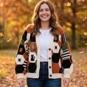 May include: A long-sleeve crochet cardigan featuring a hexagonal patchwork pattern in black, white, orange, and green. The garment includes motifs of spiders, a black cat, and pumpkins, finished with a cream-colored button-down front placket.