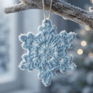 Op de afbeelding: Een lichtblauw gehaakt sneeuwvlokornament dat aan een met vorst bedekte tak hangt. De zespuntige stervormige decoratie is voorzien van ingewikkelde garenlussen en een centraal cirkelvormig patroon, opgehangen aan een dun, natuurkleurig touwtje.